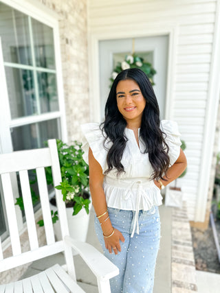 Woman standing on a porch wearing a white ruffled top and light blue jeans.