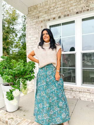 Woman in a floral skirt and beige sweater standing on a porch.