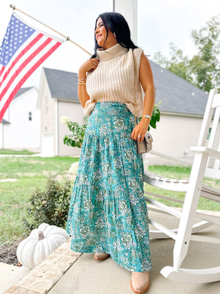 Woman in a beige sweater and blue floral skirt standing on a porch with an American flag in the background.