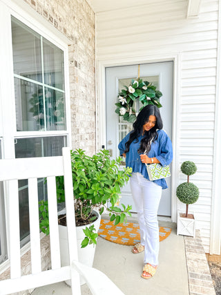 Woman standing on a porch with plants and a wreath, wearing a blue shirt and white pants.