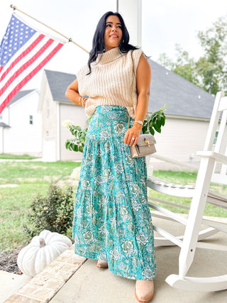 Woman in a floral skirt and beige sweater standing on a porch with an American flag in the background.