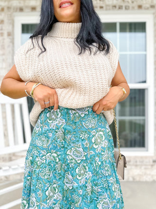 Person wearing a beige sleeveless sweater and blue floral skirt with a neutral background